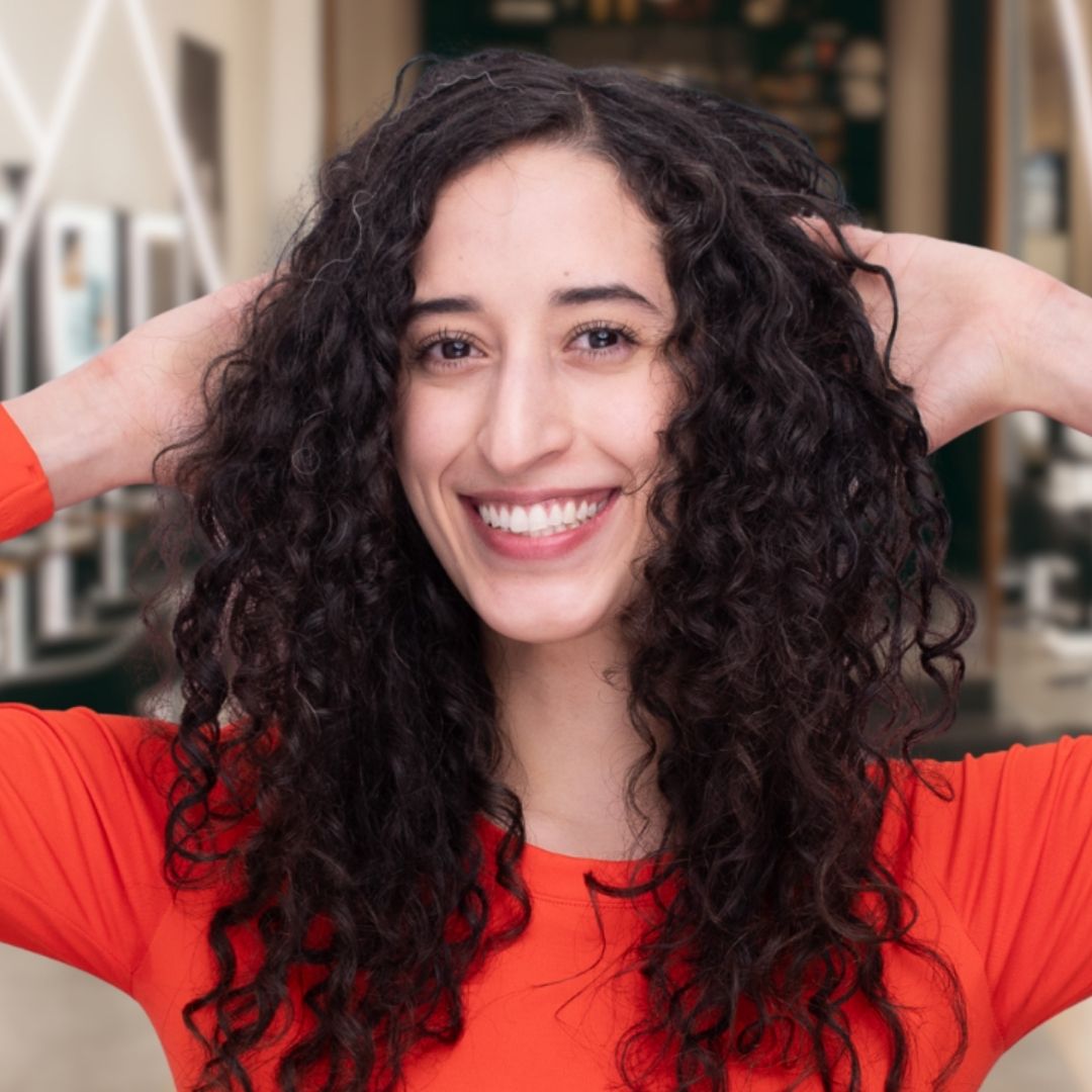 woman in red shirt with brown curly hair smiling, with her hand behind her back, showing off the volume of her hair