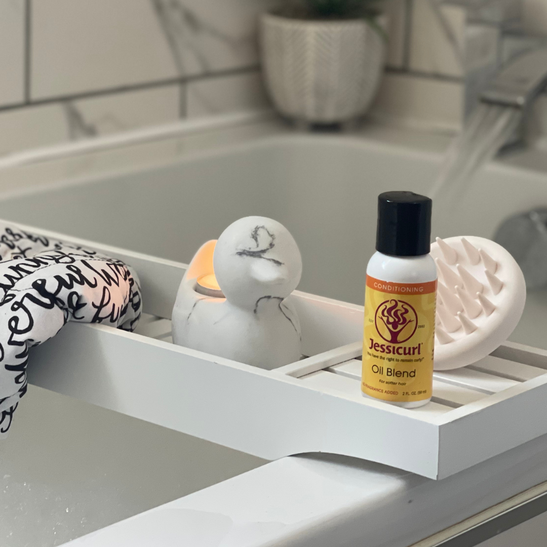 Jessicurl Oil Blend on a white bath tray next to an exfoliating scalp massager, a candle, and a Hot Head Deep Conditioning Flaxseed Cap