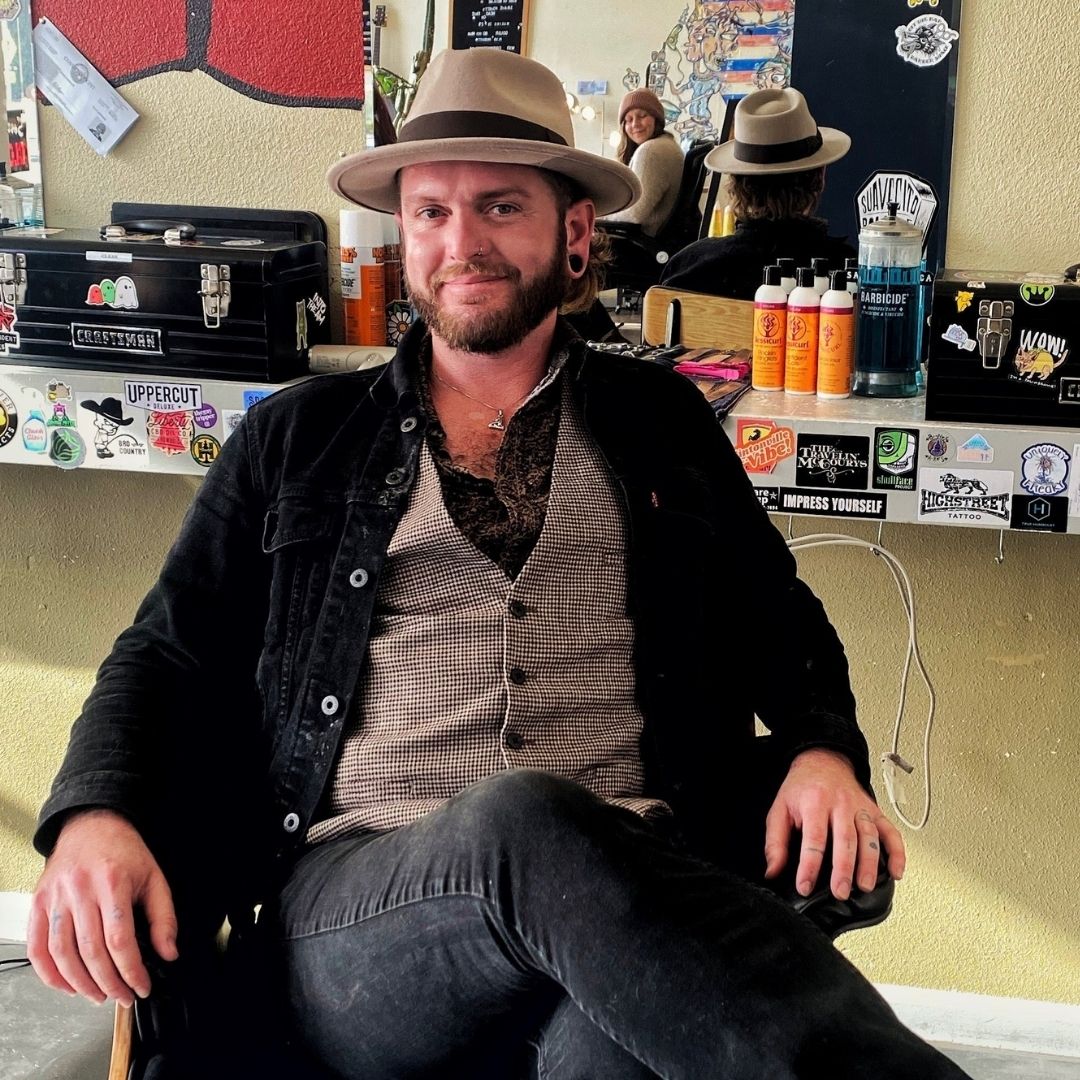 Curly haired male barber in a hat and casual suit sitting it his own chair at his work station