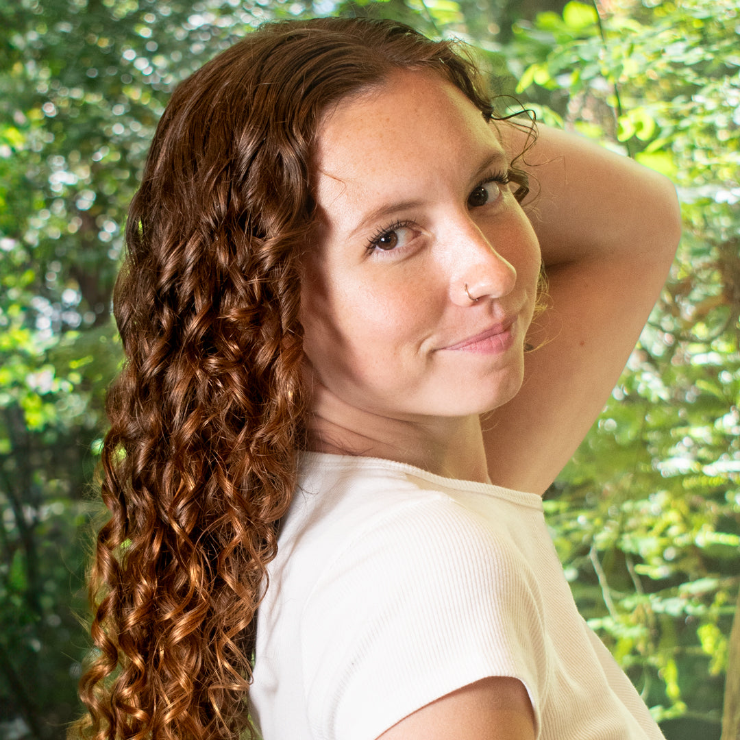 smiling curly haired woman looking at the camera from the side with trees behind her