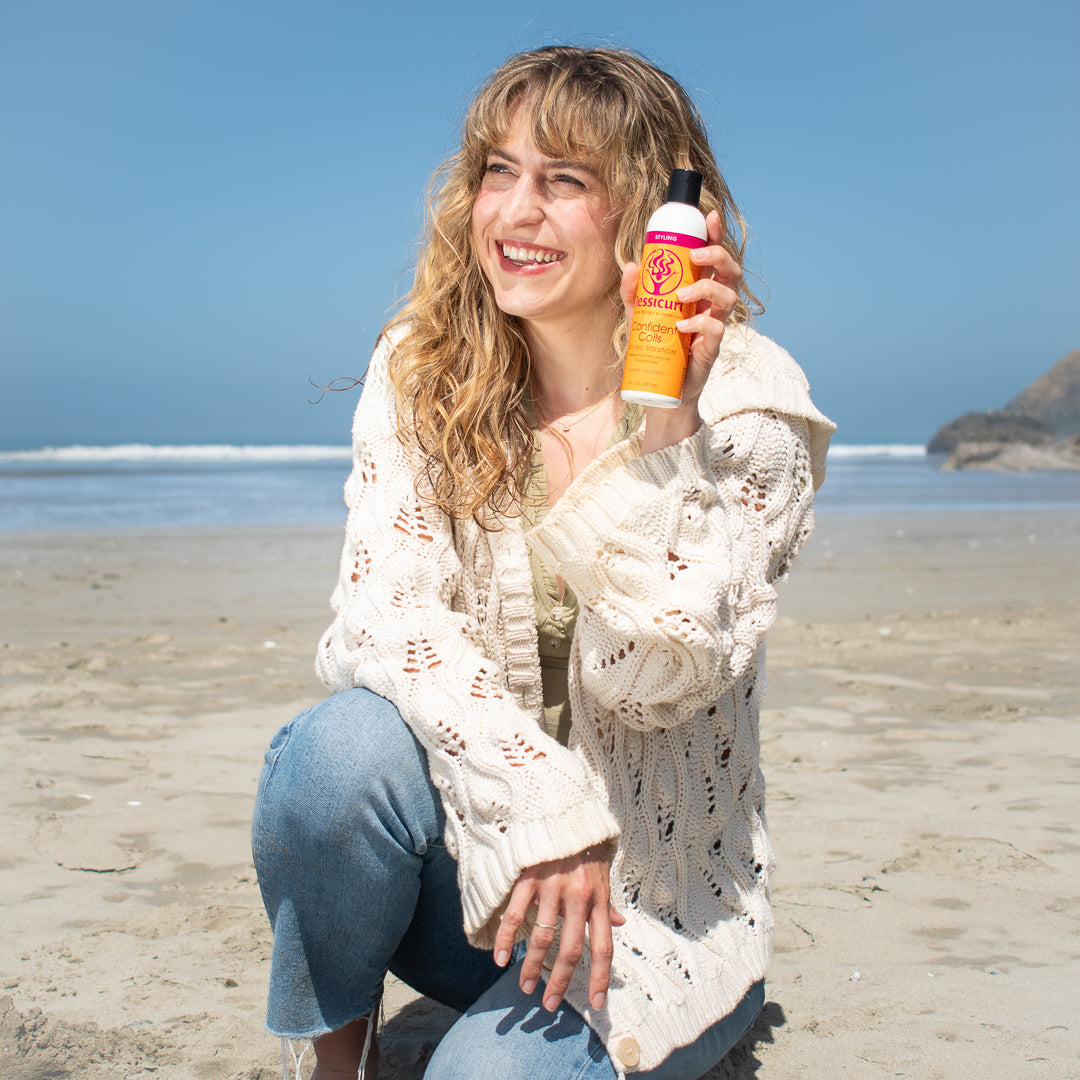 curly haired woman on the beach kneeling and holding up a bottle of Confident Coils Styling Solution