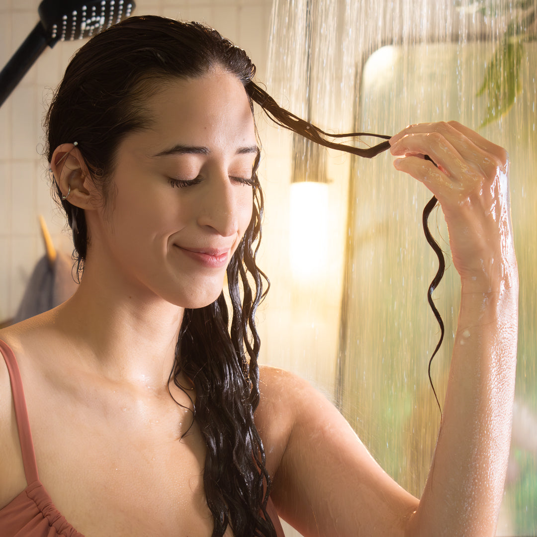 curly haired woman in the shower, smiling while holding out a curl