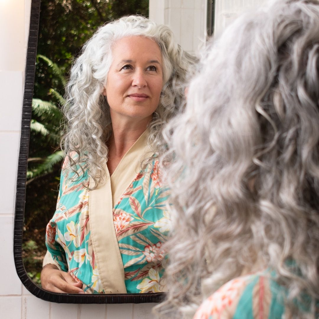 woman with long, gray, curly hair, smiling and looking in a bathroom mirror