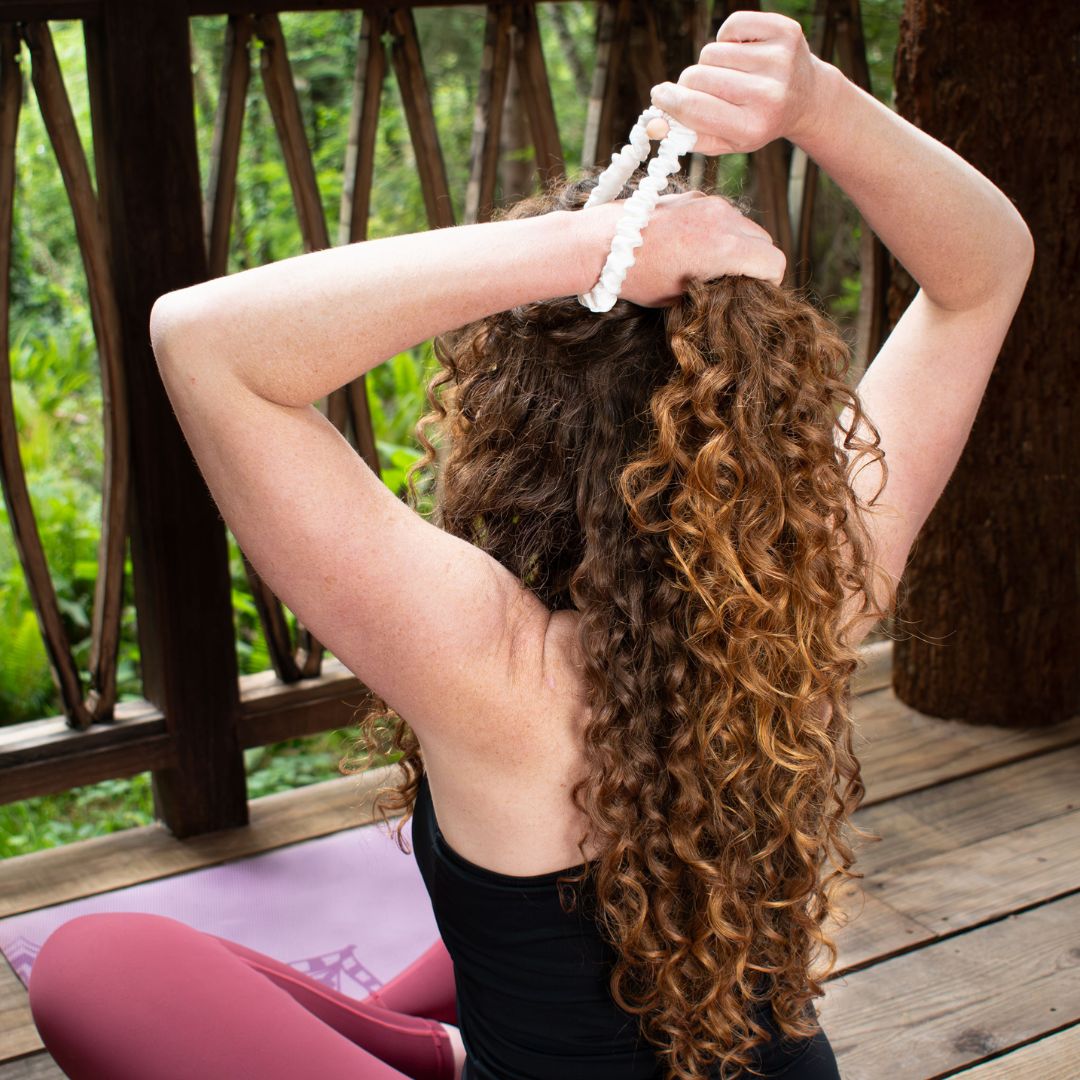 curly haired woman sitting on a porch in the woods putting her hair up with a scrunchie