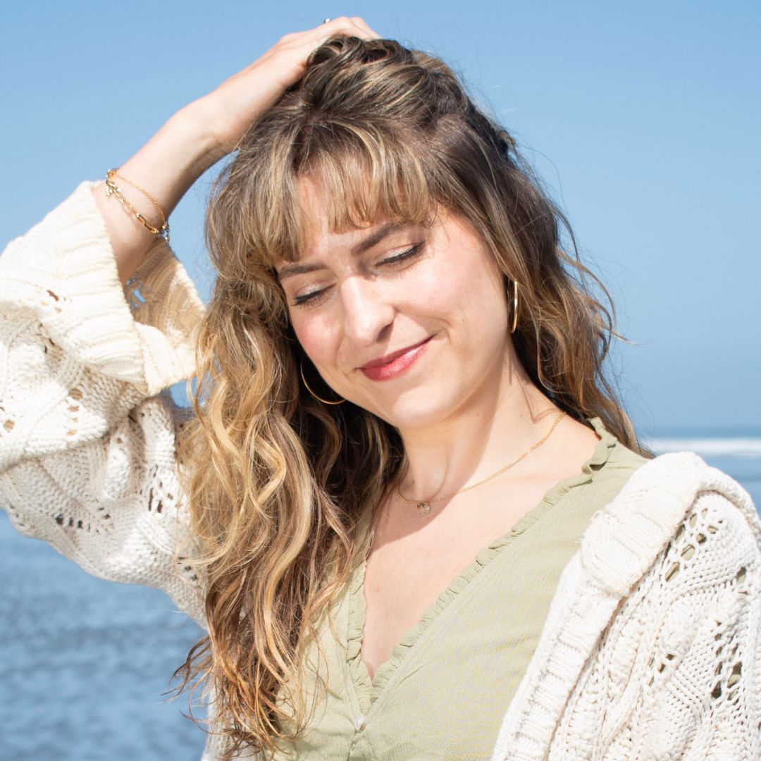 Girl with wavy hair brushing it back with her fingers, smiling downwards with the ocean behind her.