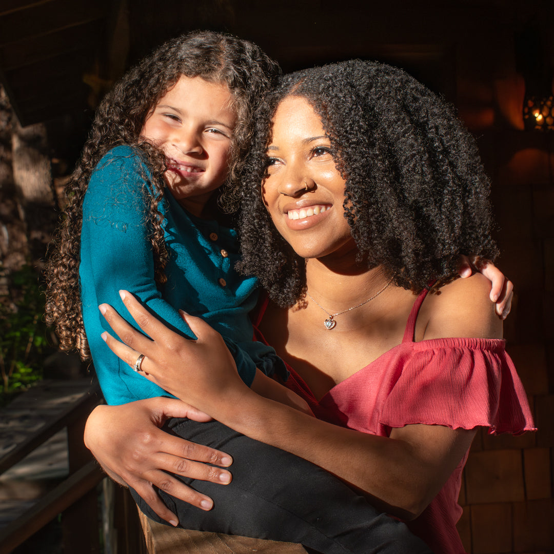 Curly haired mother and daughter sitting in the sun, smiling and holding each other