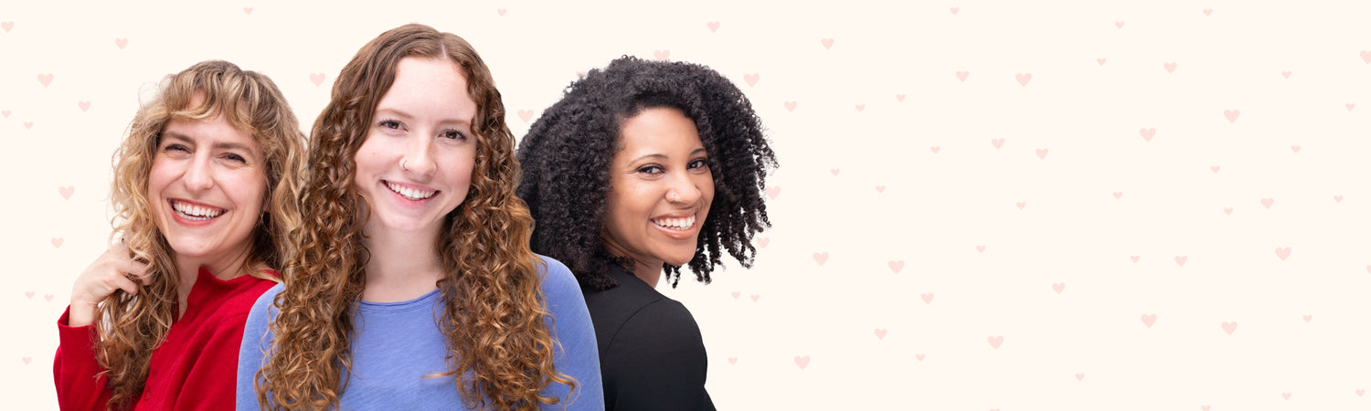 Three women (one with wavy, one with curly, and one with coily hair) smiling and multiple subtle little hearts in the background.